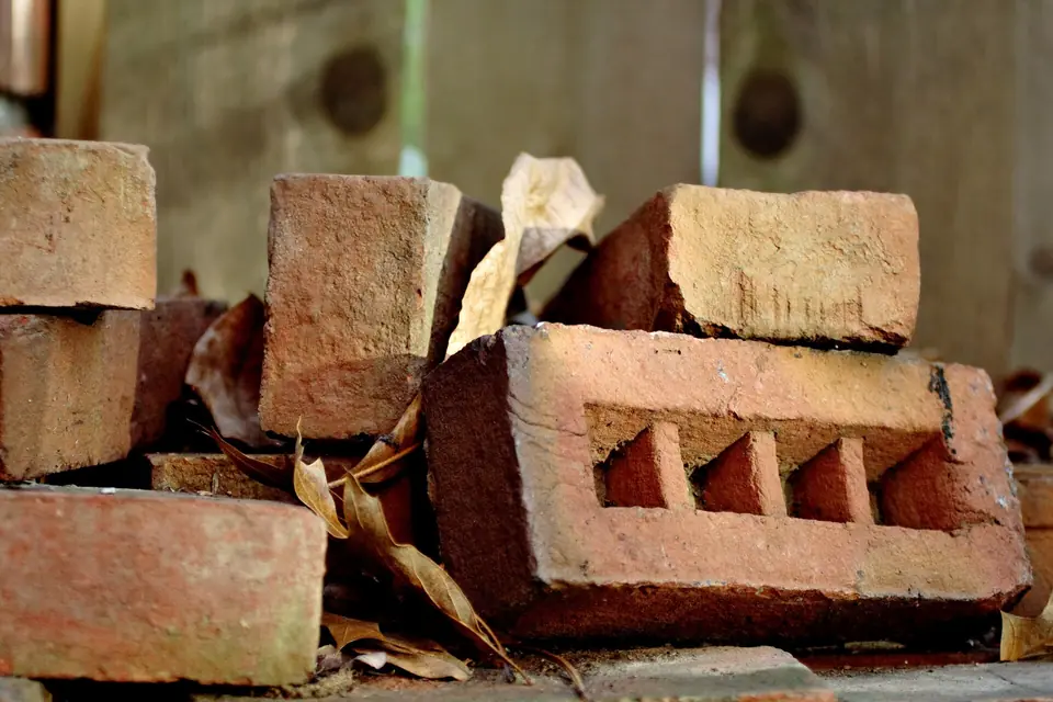 Tas de briques en terre cuite, dont une brique perforée, empilées sur un chantier parmi quelques feuilles mortes.